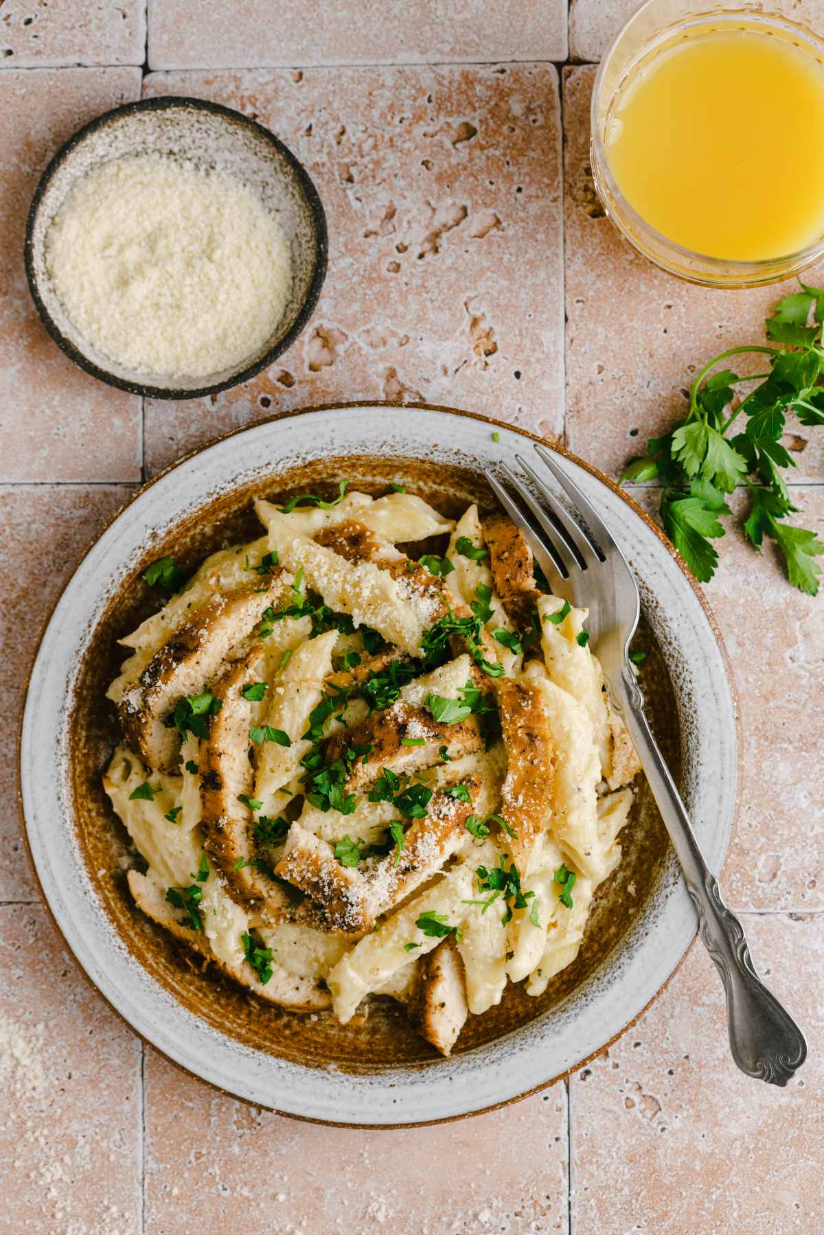 An overhead shot of creamy garlic chicken pasta with a small bowl of parmesan cheese and a glass of orange juice.