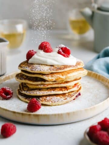 A stack of pancakes with fruit and cream sauce on a plate, being dusted with powdered sugar.