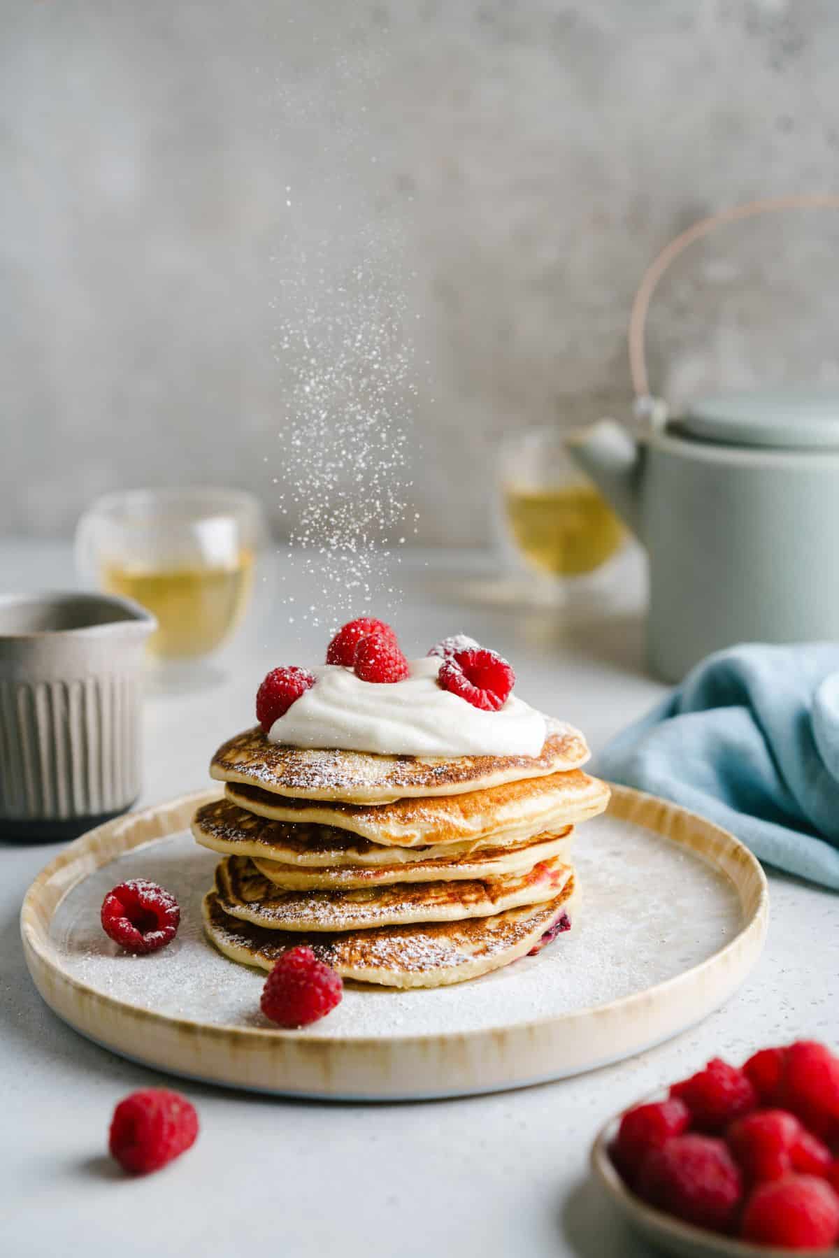 A stack of pancakes with fruit and cream sauce on a plate, being dusted with powdered sugar.